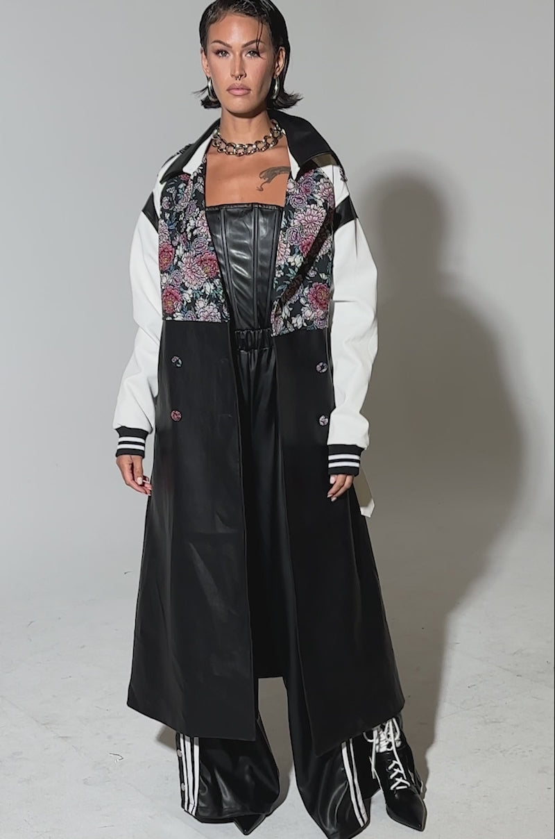 Video of woman modeling the END GAME MIXED MEDIA TRENCH, a long black and white coat with a floral-patterned upper section, paired with black leather pants, lace-up boots, and a chunky chain necklace, standing against a plain background.