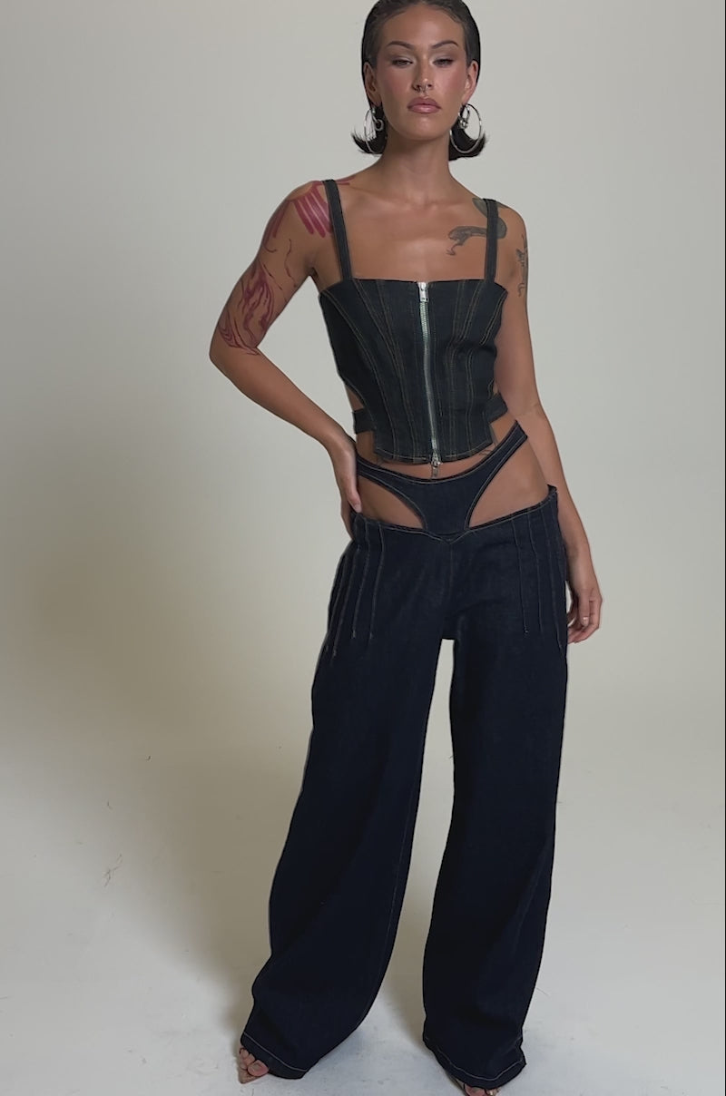 A video of a woman in the SEXY CAN I RELAXED DENIM, a Y2K-inspired corset-style zip-up top paired with low-rise wide-leg jeans featuring a bold cut-out waistband. She has wet-look hair and visible arm tattoos.