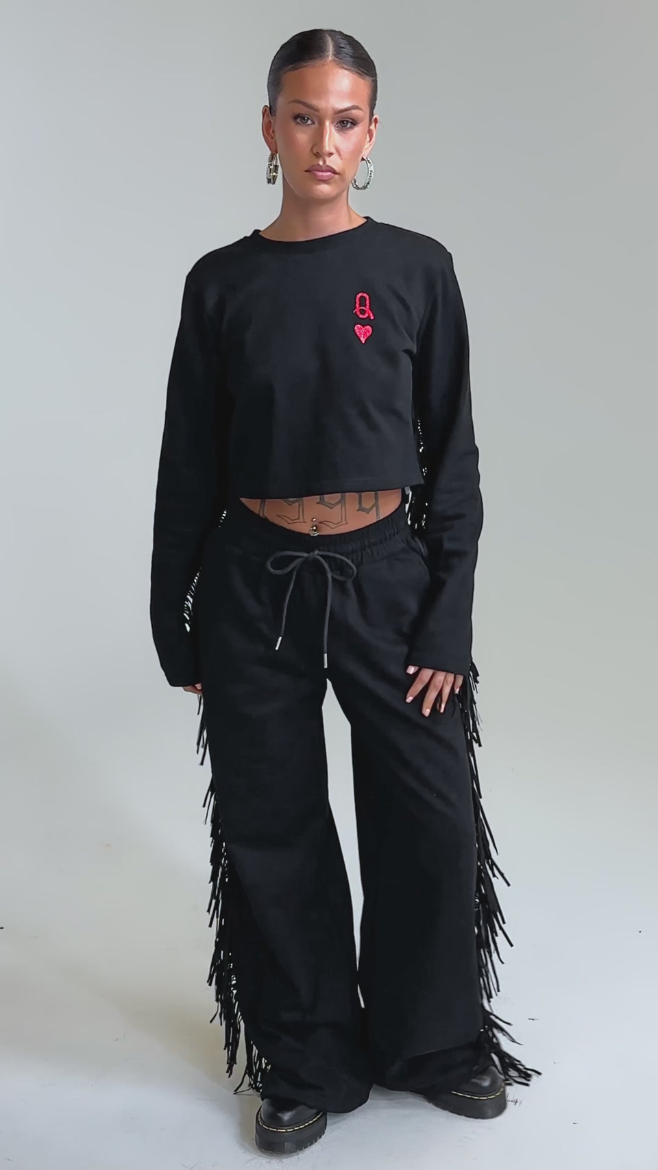 Video of woman modeling the PLAYING CARDS WIDE LEG JOGGER and a matching long-sleeve crop top, her hands in her pockets. Hoop earrings and black shoes complete her look, highlighting the stylish fringe detailing down the jogger’s sides.