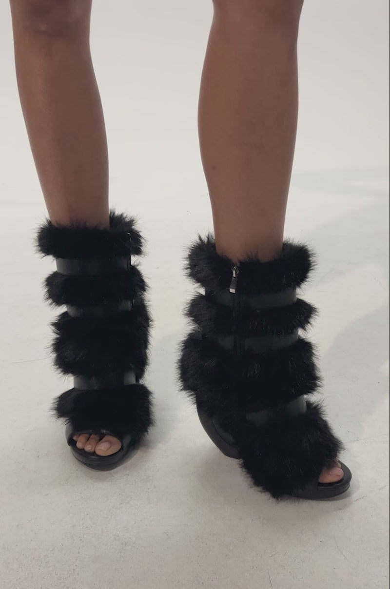 Video of woman modeling the AZALEA WANG ELIANA BLACK FUR OPEN TOE WEDGE BOOTIE, featuring multiple thick faux fur straps and a unique curved wedge heel, shown from the calves down against a plain background