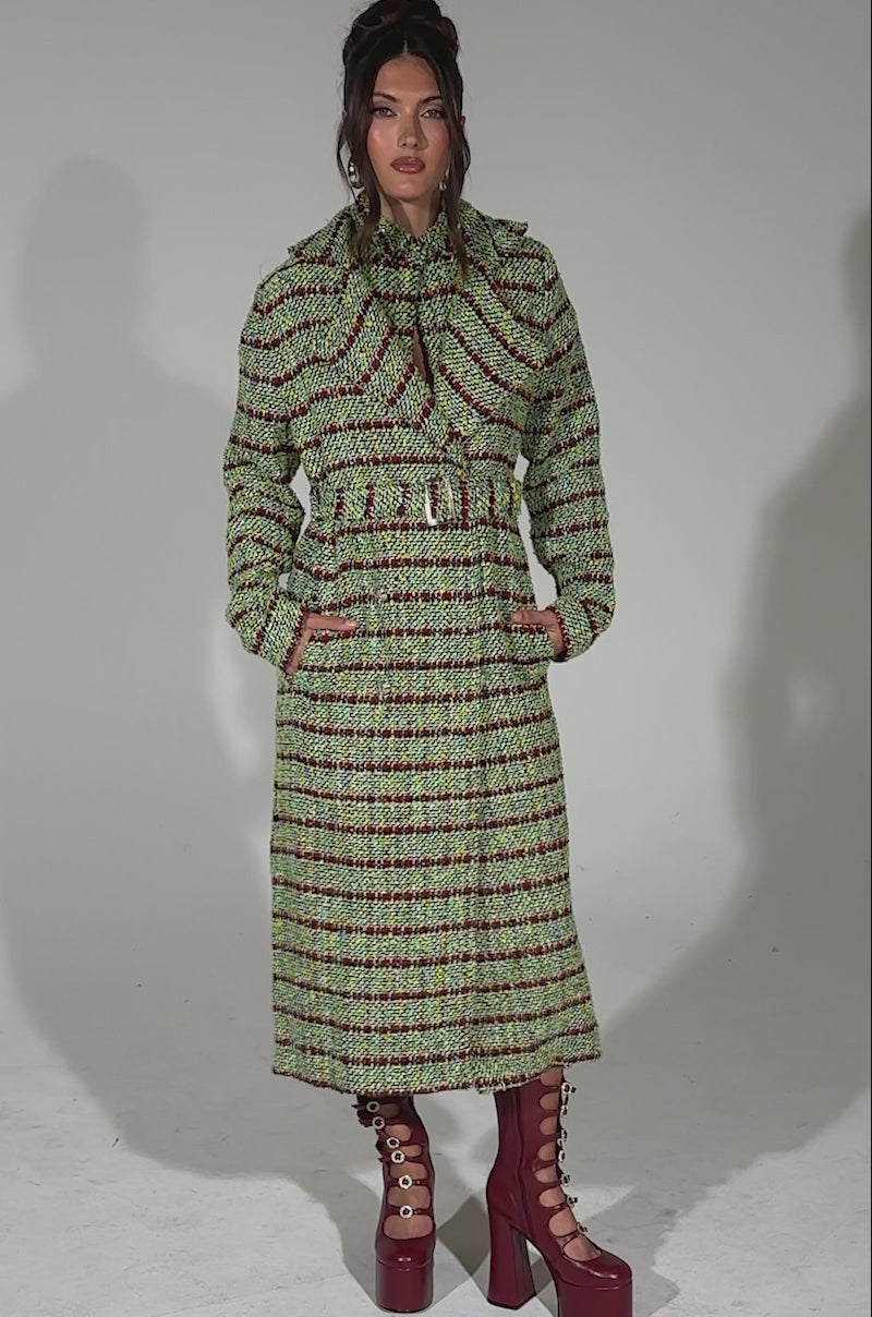 A video of a woman wearing the IT'S GIVING LUCK TWEED TRENCH, a longline multicolor tweed coat with green, red, and black stripes. She styles it with maroon platform boots and a matching scarf, posing against a light blue background.
