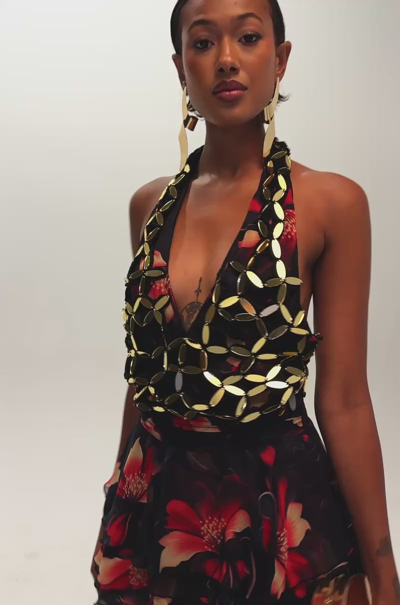Video of a woman modeling the ISLAND TIME EMBELLISHED MINI DRESS, which features a sleeveless black halter top with gold accents and a black ruffled skirt adorned with red floral patterns.