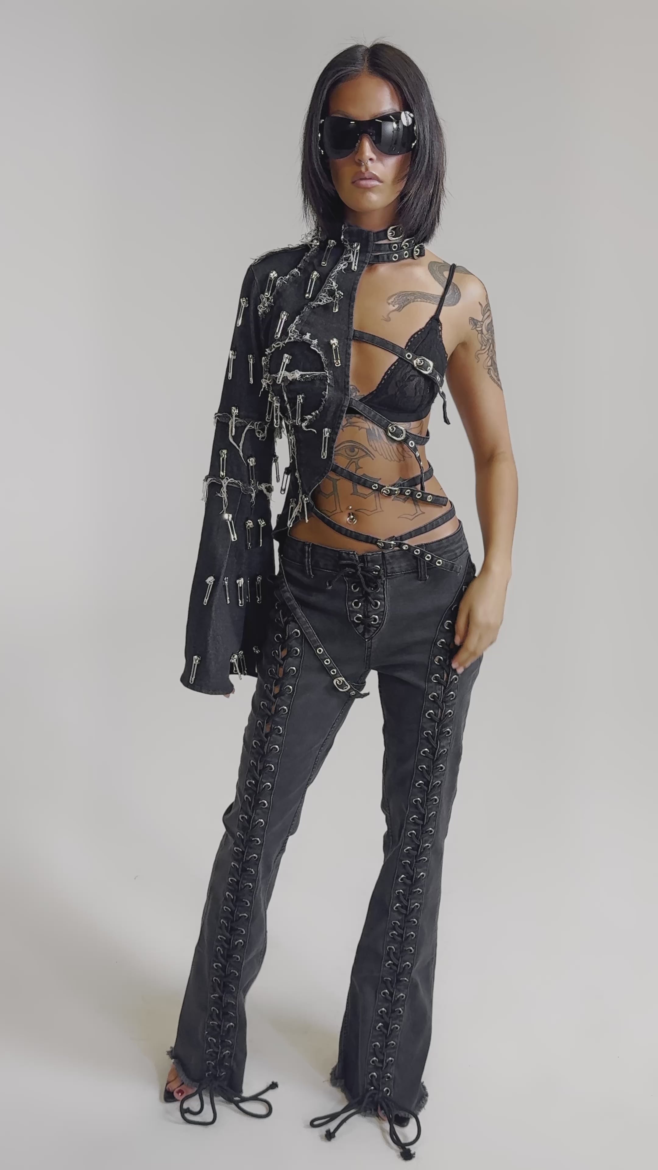 A video of a woman wearing the HOTTY LACE UP FLARE DENIM pants with a matching asymmetrical jacket featuring metal details, a black bra, and sunglasses.