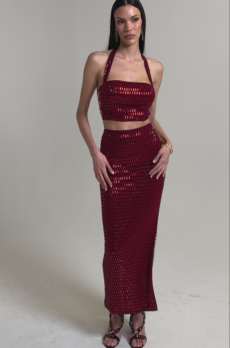 A video of a woman in the FALLING IN LOVE RHINESTONE MAXI SKIRT, a high-rise burgundy embellished maxi paired with a red halter crop top, strappy heeled sandals, and sleek straight hair.