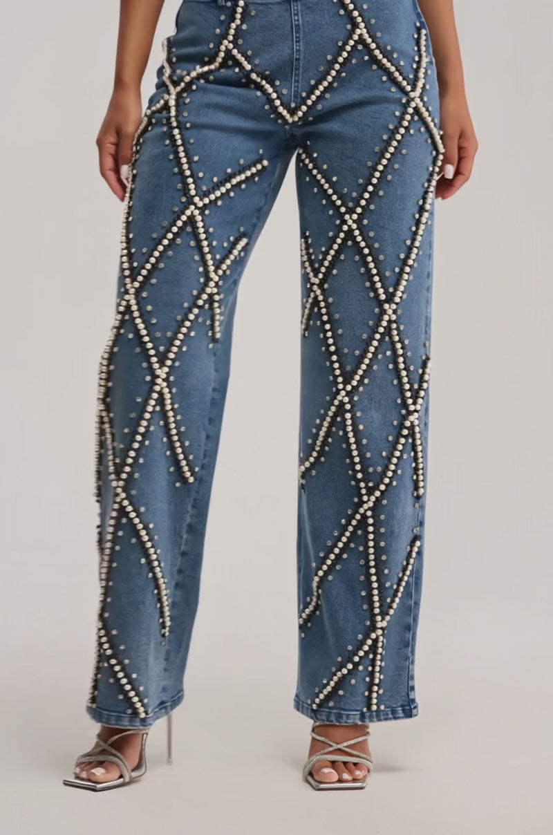 Video of woman modeling the DRIPPING IN PEARLS DENIM PANT—high-waisted, wide-leg jeans detailed with crisscrossing pearl embellishments—styled with silver high-heeled sandals for a chic, statement look.