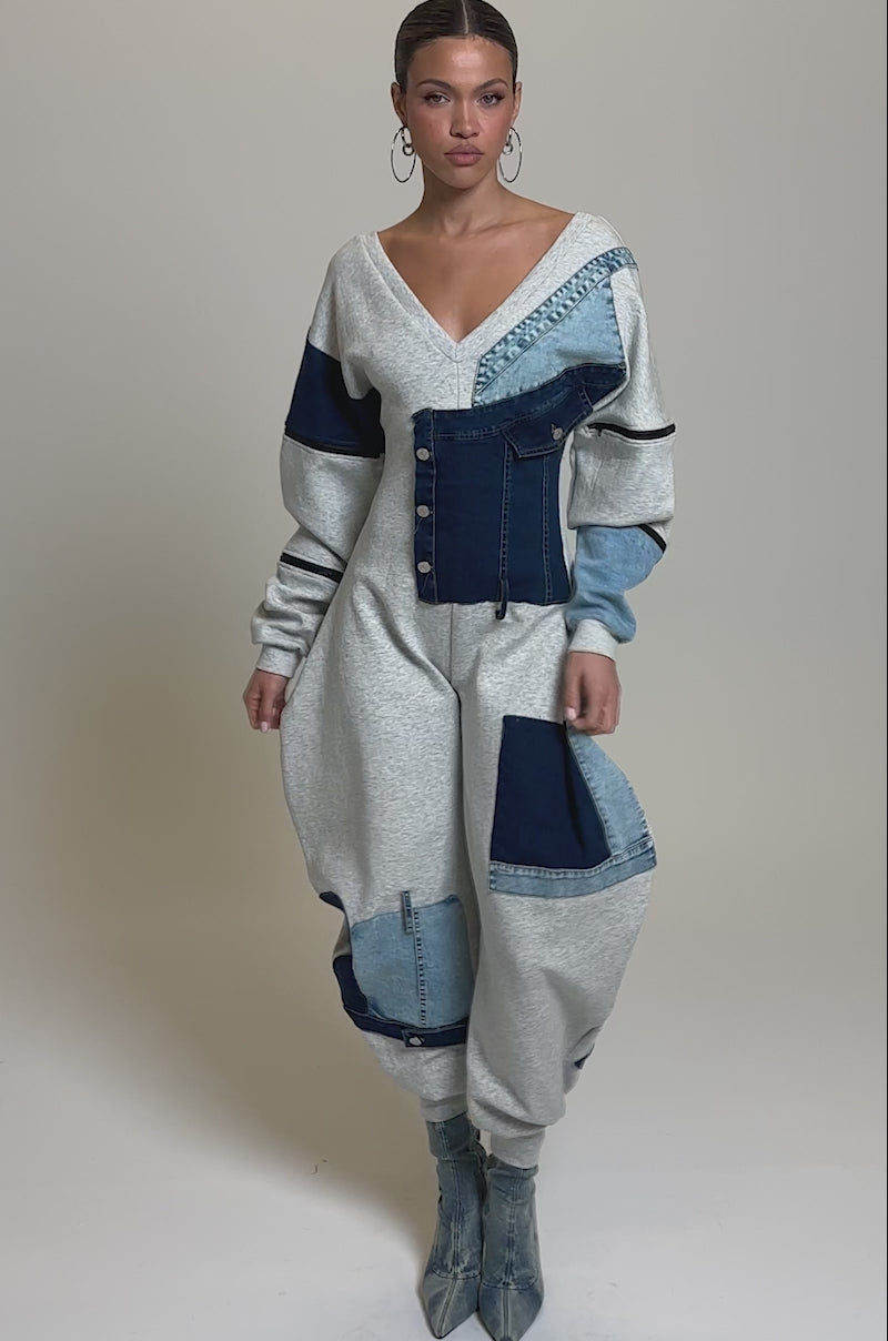 Video of woman modeling the COMFY COZY SWEATSHIRT JUMPSUIT IN PATCHWORK DENIM, featuring a V-neckline and loose-fitting pants. She styles it with denim ankle boots and large hoop earrings against a plain background.