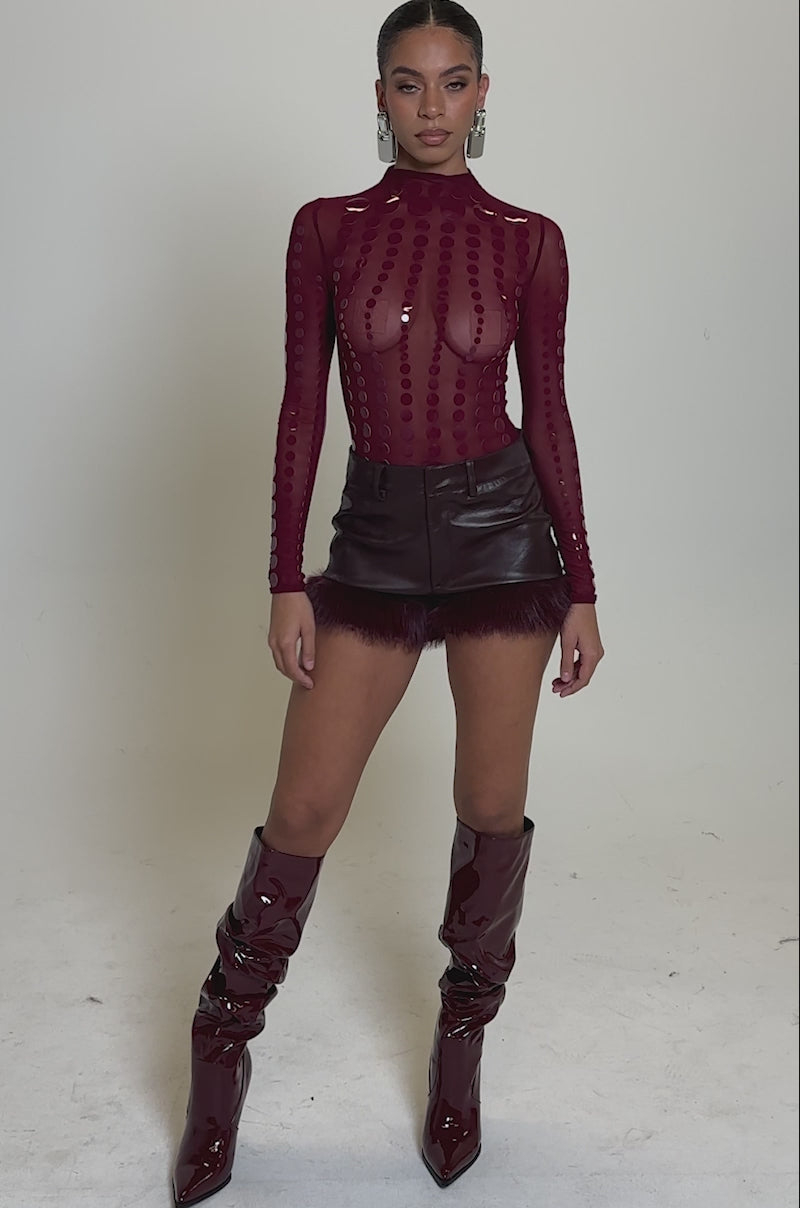 A video of a woman wearing an AKIRA Label TRIPPY TYPE BODYSUIT, high-waisted maroon leather shorts with feather trim, and slouchy, shiny maroon knee-high boots.