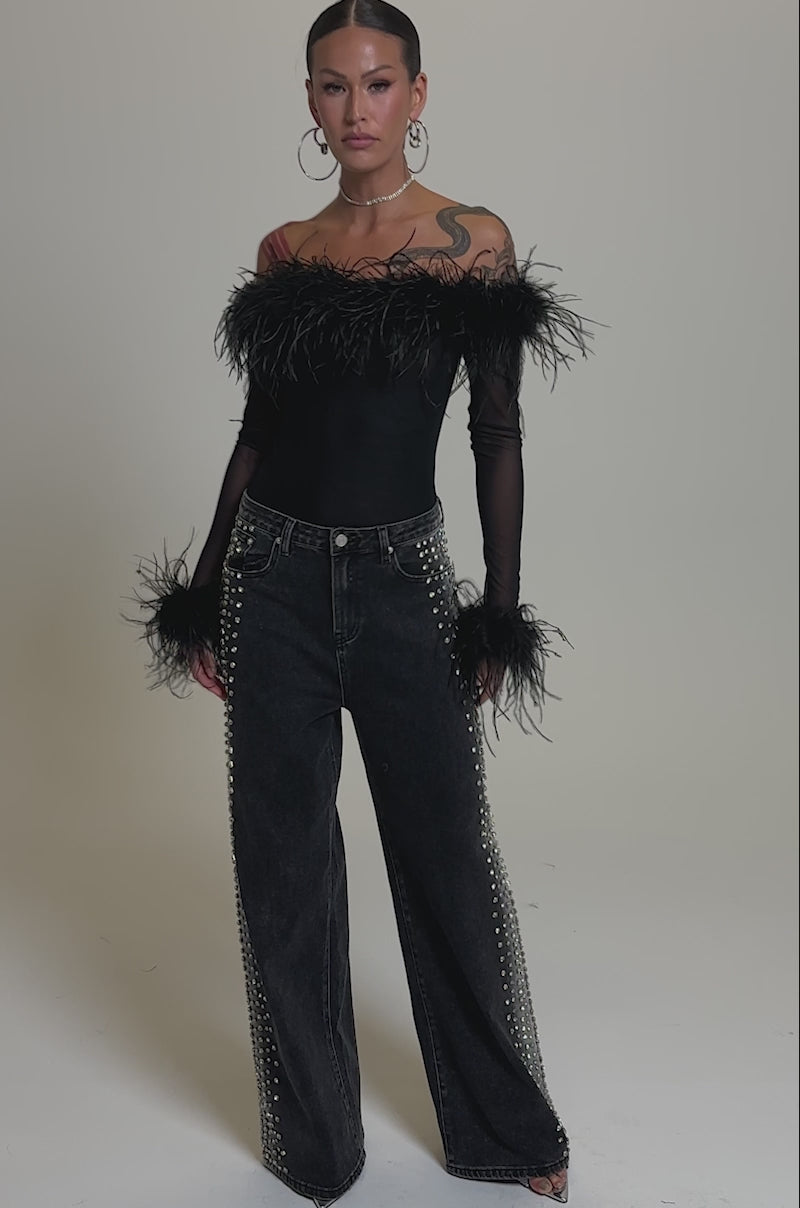 A video of a woman in a sheer black off-shoulder top with feather details, paired with FREAKING FIRE EMBELLISHED DENIM.