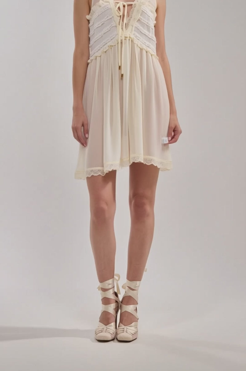 Video of woman modeling the FREE SPIRIT LACE MINI DRESS, an ivory, sleeveless mini dress with ruffles and lace details finished with a bow at the neckline, paired with ballet-inspired lace-up shoes.