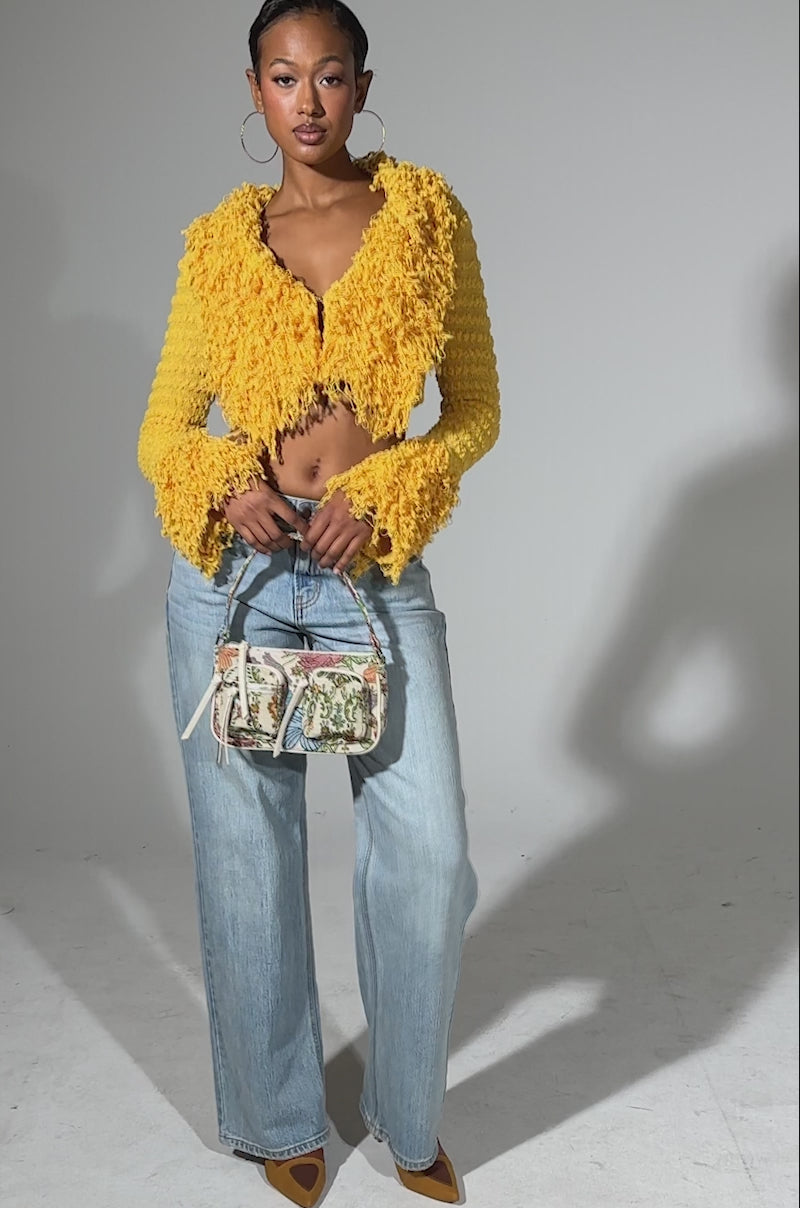 A video of a woman standing confidently in the SUNSHINE QUEEN FRINGE SWEATER, paired with light blue wide-leg jeans, yellow heels, large hoop earrings, and a small floral handbag against a light background.