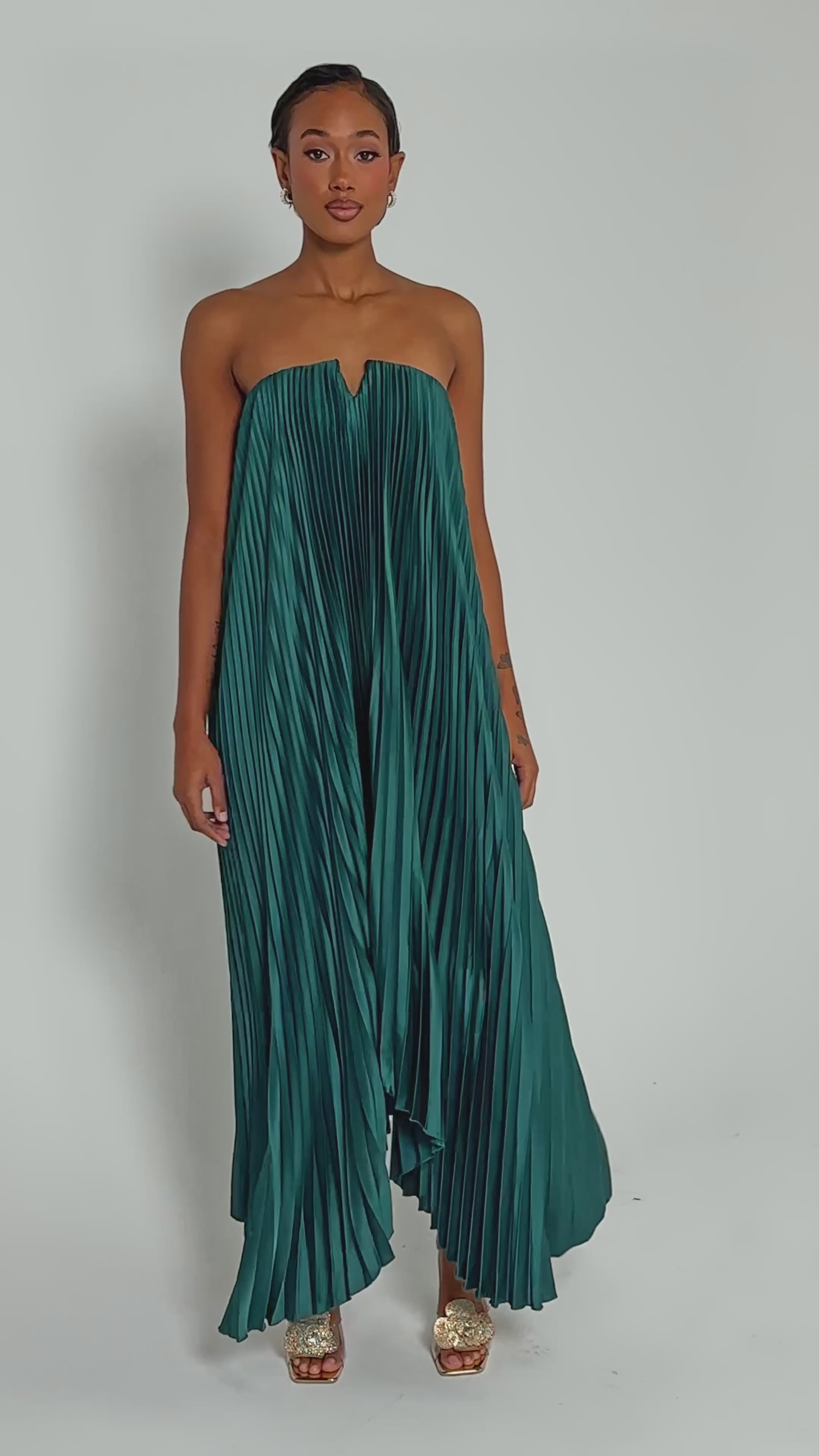 Video of woman modeling the EMERALD DREAM SLEEVELESS MAXI DRESS in emerald green satin with pleats, paired with gold open-toed heels, against a plain light background.