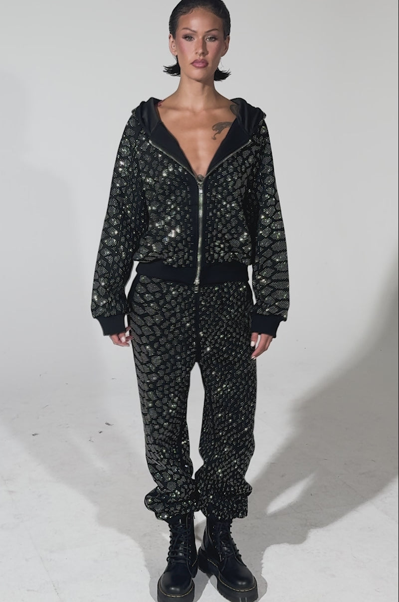 A video of a woman standing wearing the SNEAKY LITTLE SNAKE WIDE LEG JOGGER, featuring a black quilted design with a shiny silver geometric pattern, paired with a matching jacket and black lace-up boots against a plain background.