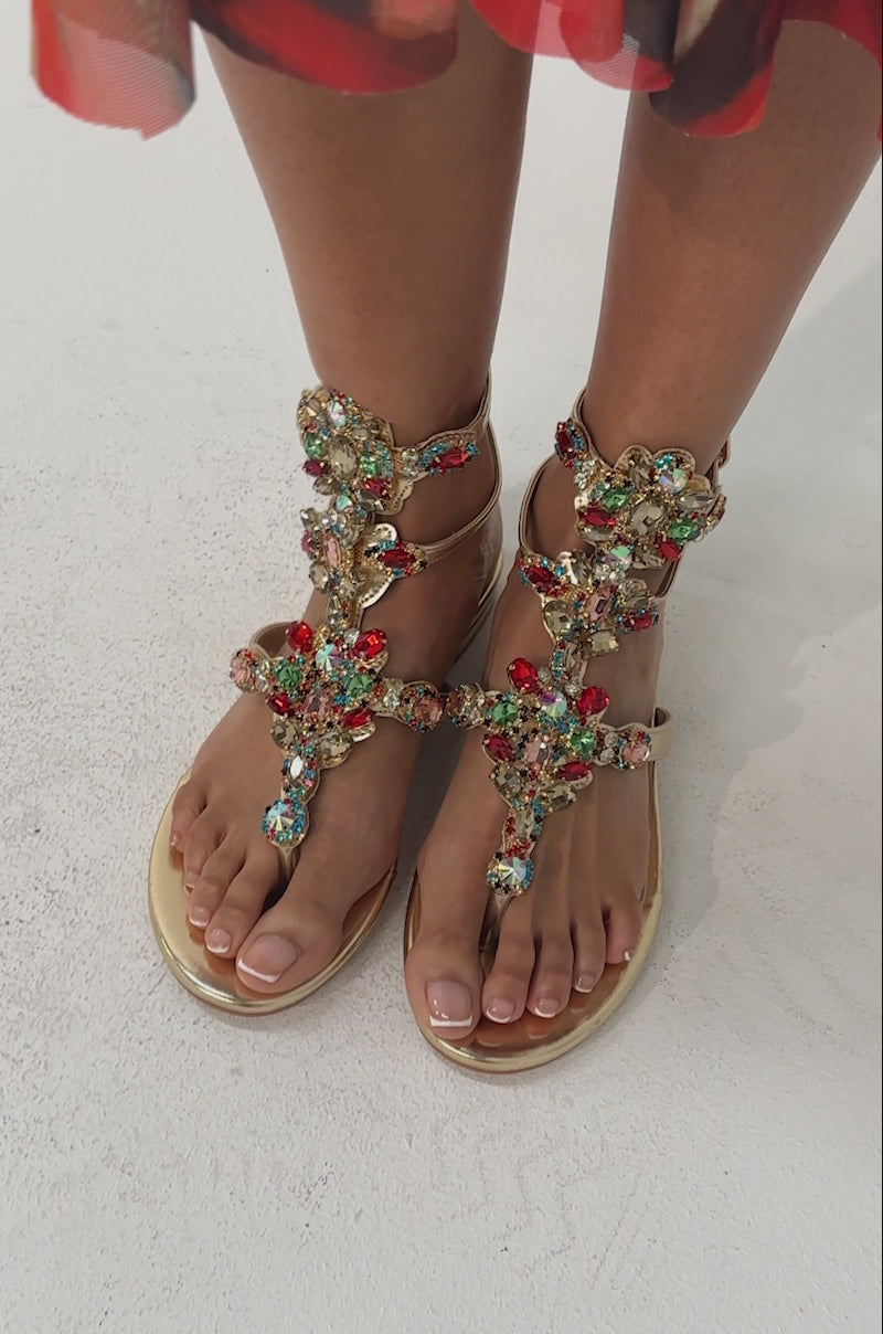 Video of woman modeling the AZALEA WANG OLISA GOLD RHINESTONE EMBELLISHED FLAT SANDALS paired with a vibrant red and orange patterned skirt, with the photo highlighting their feet and lower legs.