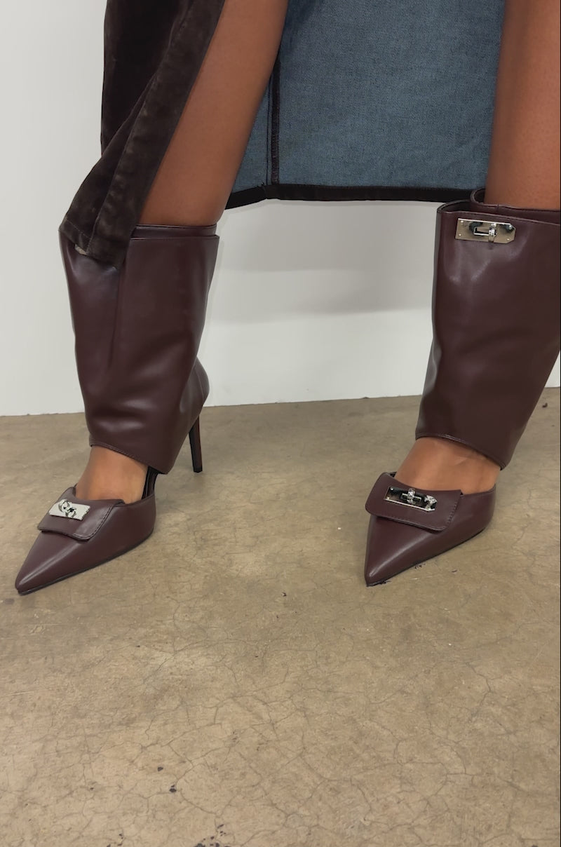 A video of a model wearing a dark skirt with a high slit, someone showcases bare legs and the AZALEA WANG SARVESH BROWN CUTOUT STILETTO BOOTIE while standing on a concrete floor against a plain background.