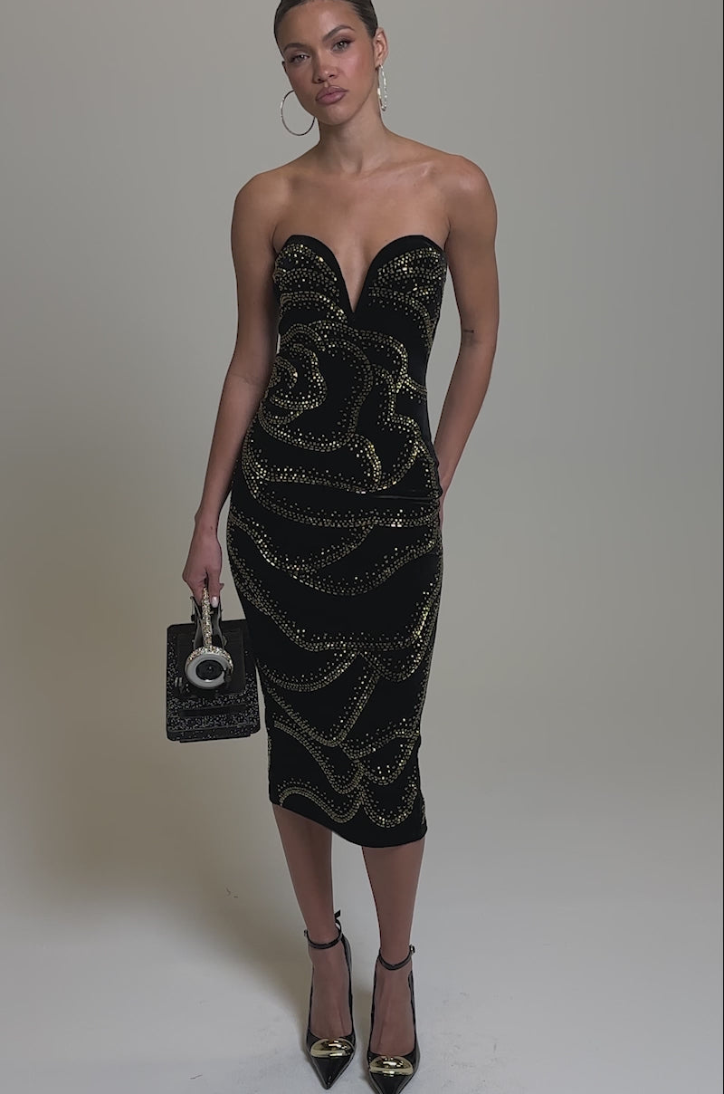 Video of woman modeling the SHIMMER IN GOLD MIDI DRESS, a strapless velvet piece with gold accents. She holds a black sewing machine-shaped handbag, wears hoop earrings and black pointed heels, set against a neutral background.