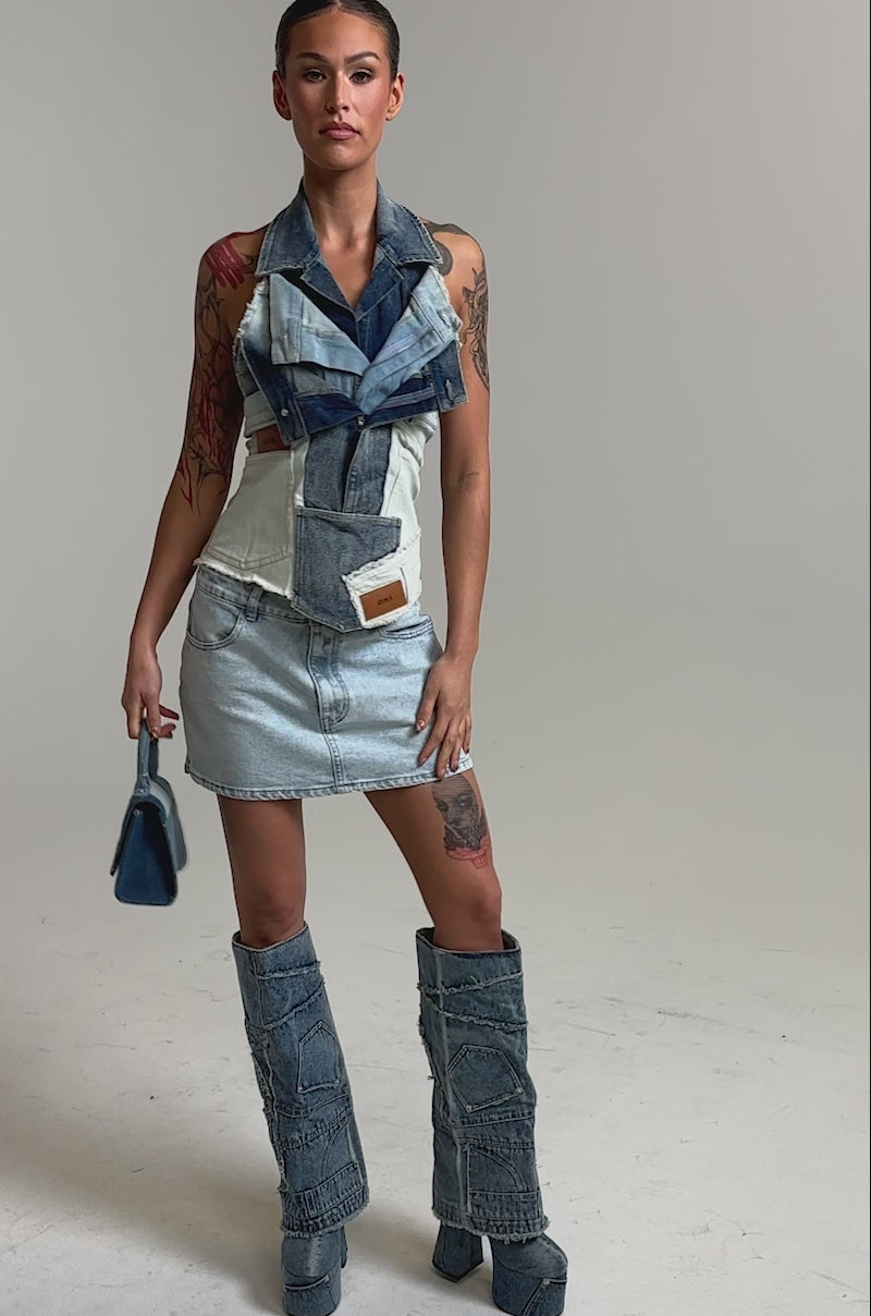 Video of woman modeling the ALWAYS RIGHT DENIM ABSTRACT BLOUSE—a sleeveless, patchwork denim top with zippers, a collar, and a corset-style silhouette in various blue tones. Her hair is pulled back as she stands against a plain light background.