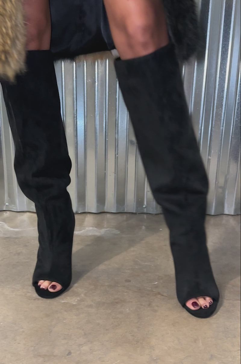 A video of a model wearing AZALEA WANG VINCENT BLACK FAUX SUEDE OPEN TOE BOOTS, black leather shorts, and a multicolored faux fur coat, a person with a thigh tattoo stands in front of a corrugated metal wall on concrete.