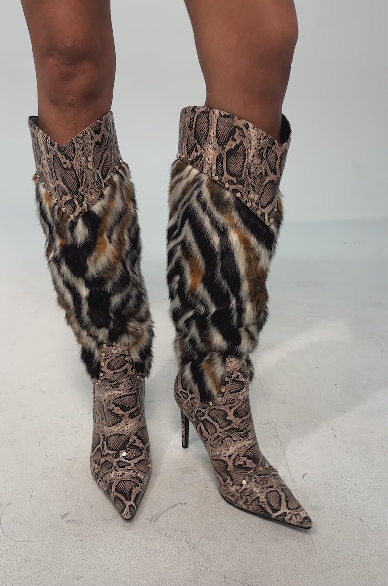 A video of a model wearing the AZALEA WANG ALISETTE SNAKE MULTI FUR STILETTO BOOT. The boot is a bold knee-high, animal print stiletto with a pointed toe, snakeskin pattern, faux fur panel, metal ring accent, and side studs.