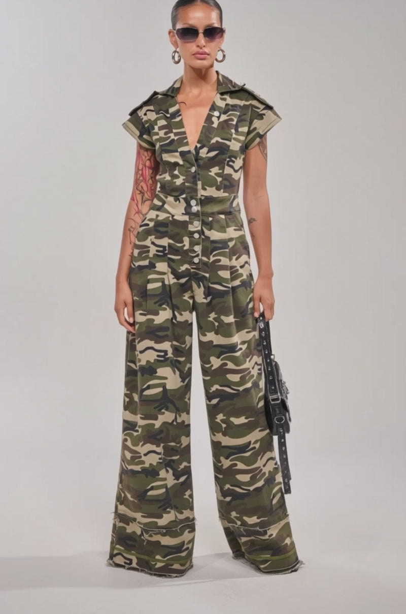 Video of woman modeling the MAJOR DRAMA CAMO JUMPSUIT IN CAMO, styled with sunglasses, earrings, and a black handbag