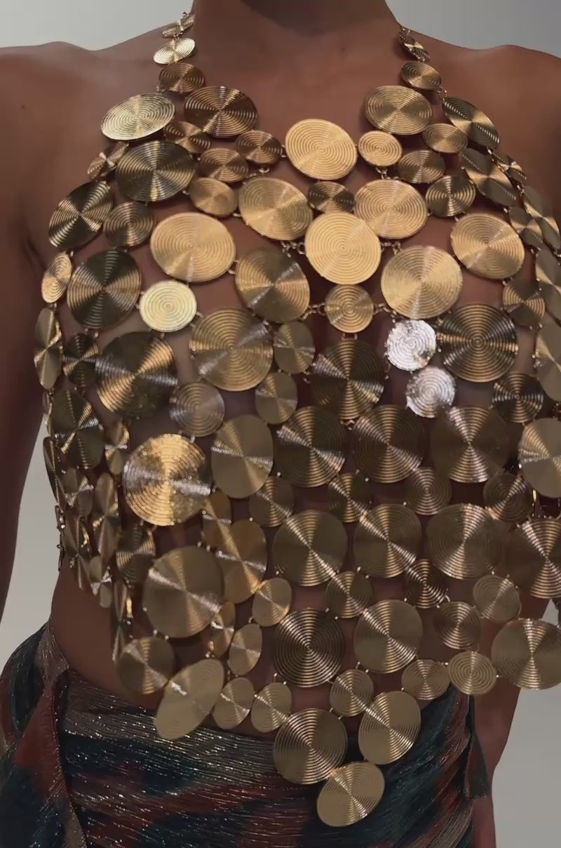 A video of a woman with dark hair in a slicked-back bun poses against a white background, wearing the GOING IN CIRCLES METAL CHAIN TOP—a halter style constructed from layered, textured gold-toned metal discs—paired with a colorful, metallic ruched midi skirt and large gold hoop earrings.