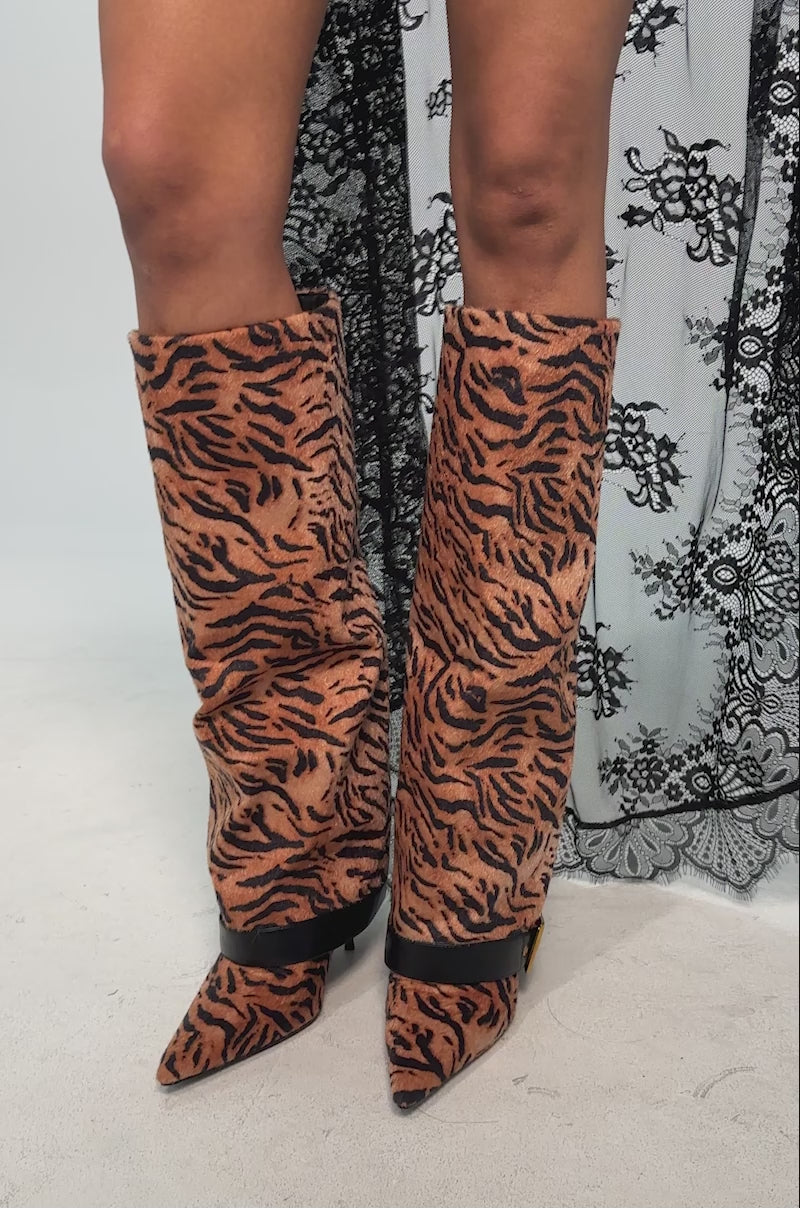 A video of a model wearing the AZALEA WANG JUST TIGER STILETTO FOLD OVER BOOT which has a knee-high, bold tiger print design with a square toe, black sole, and a black ankle strap accented by a gold buckle.