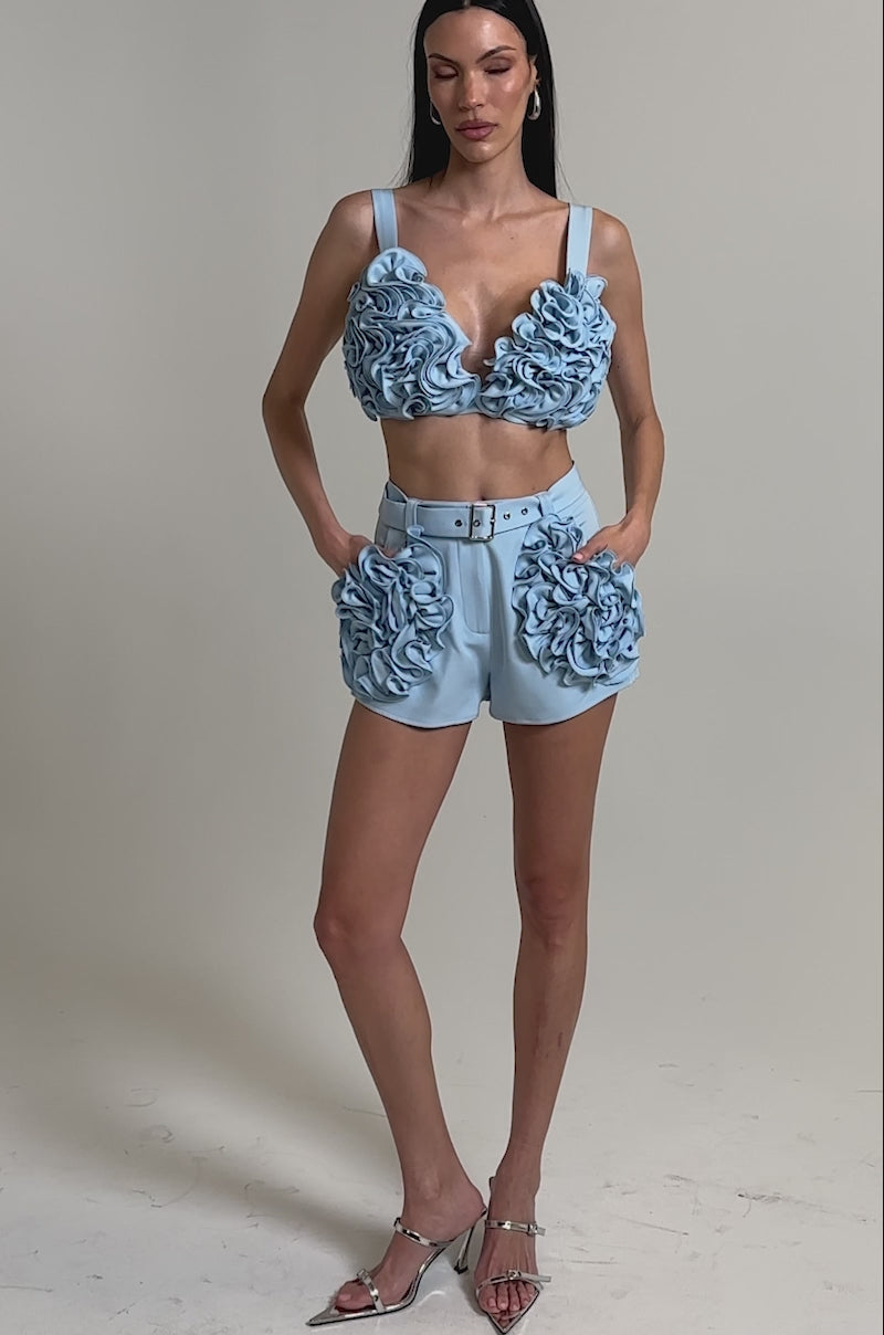 A video of a woman wearing the FLOWER GIRL BRALETTE in baby blue with large 3D floral appliqué, paired with matching high-waisted shorts. 