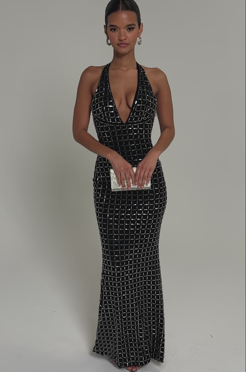 Video of woman modeling the DRESSED UP WITH NOWHERE TO GO RHINESTONE MAXI DRESS, a sleeveless, floor-length black gown with a plunging neckline and geometric rhinestone details. She holds a silver clutch, wears silver sandals, and her hair is slicked back.