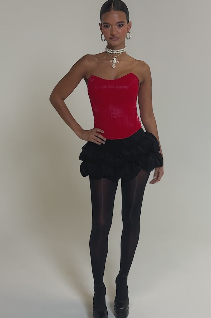 Video of woman modeling the STAY OUT MY WAY CORSET IN RED, a black textured mini skirt, black tights, hoop earrings, and layered necklaces while standing against a plain light gray background.