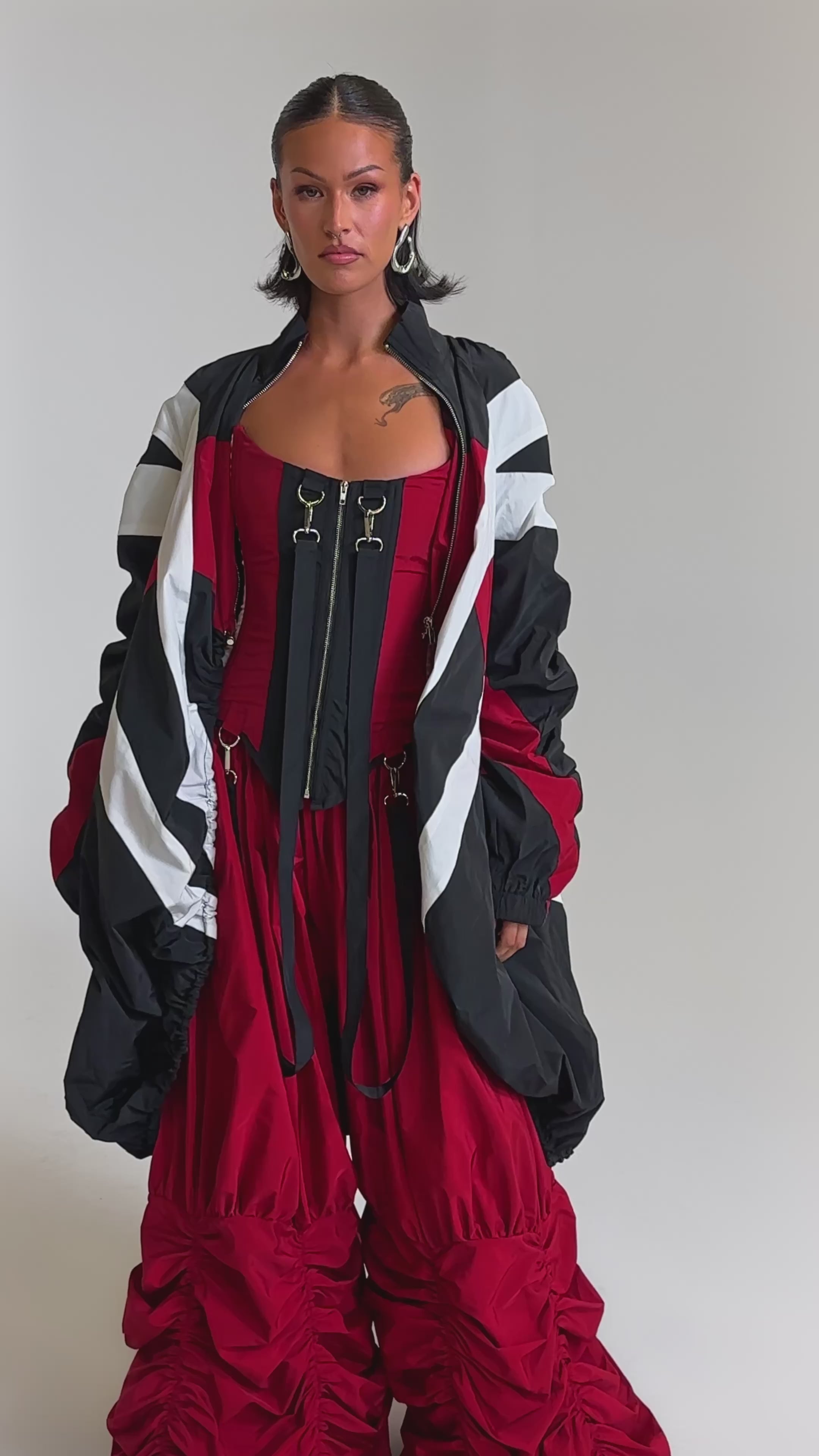 A video of a woman in the DON'T MIND ME CORSET—bold red and black—styled with a billowy red ruched skirt and an oversized black, red, and white jacket. She completes the look with slicked-back hair and neutral makeup.