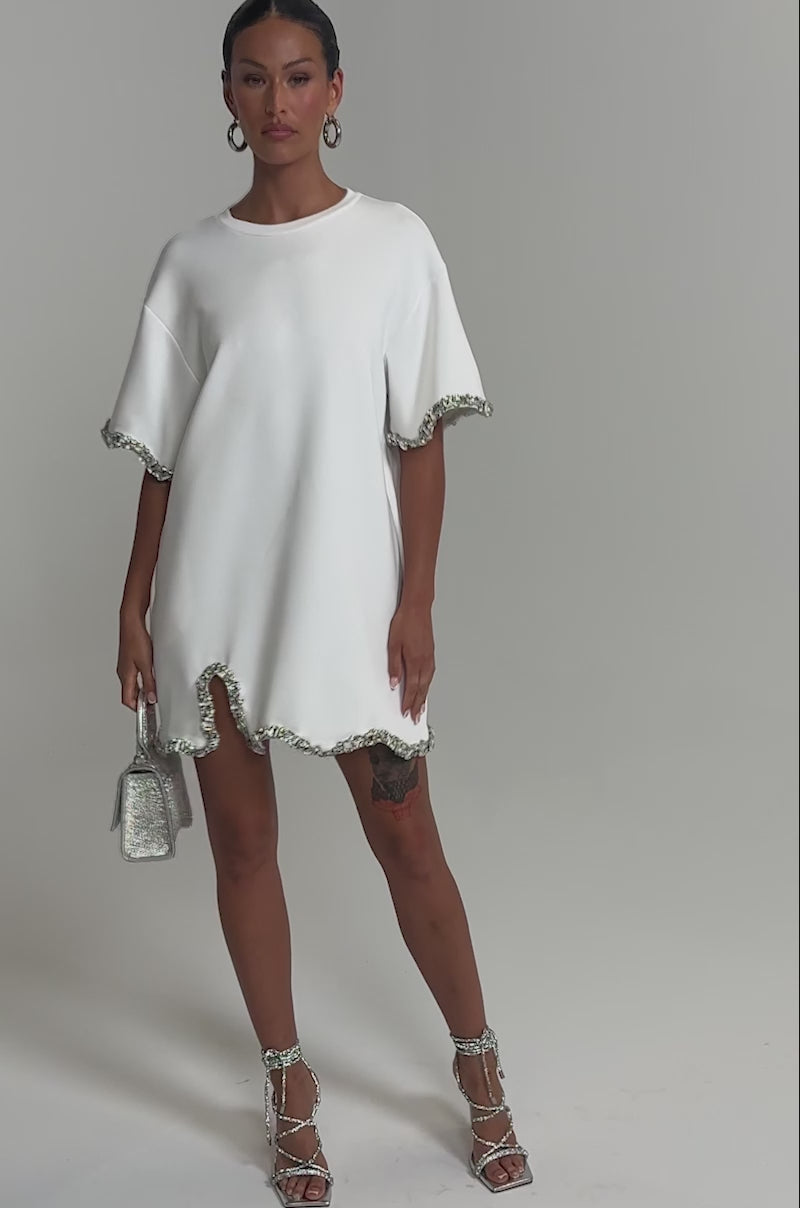 Video of woman modeling the COOL GIRL EMBELLISHED TEE SHIRT DRESS IN WHITE with jeweled trim and sleeves holding a sparkly handbag. She has hoop earrings, a sleek hairstyle, and a visible tattoo on her left thigh.