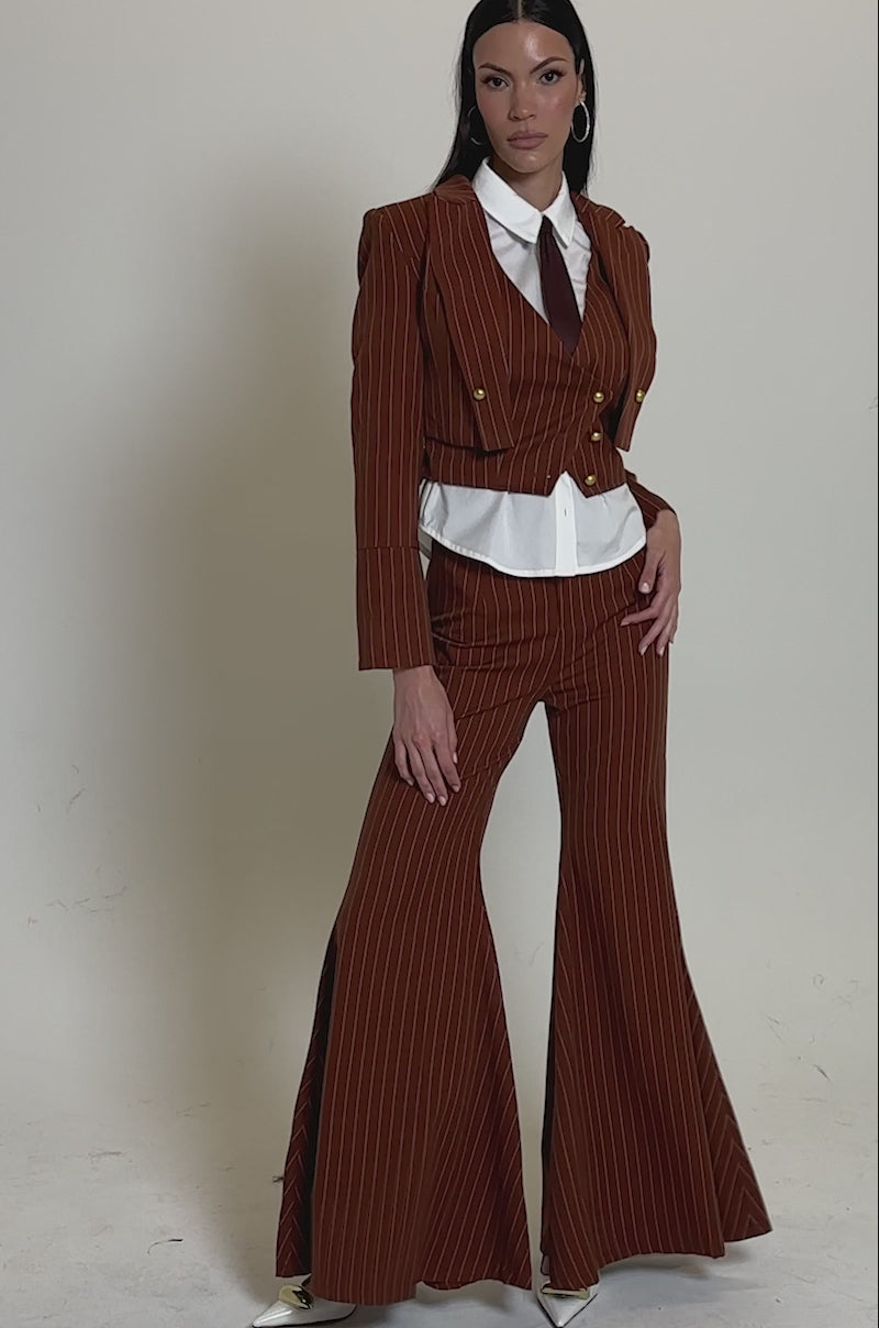 A video of a woman in the BUSINESS FIRST STRIPE BLAZER—a rust-colored cropped pinstripe blazer—styled with matching flared trousers, a white shirt, burgundy tie, and gold heels against a plain background.