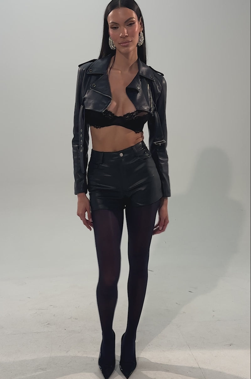 A video of a woman in the HARLEY LUXE ULTRA CROP MOTO IN NAVY, paired with a black crop top, high-waisted denim shorts with front zip, sheer black leggings, and pointed navy heels against a plain light background.