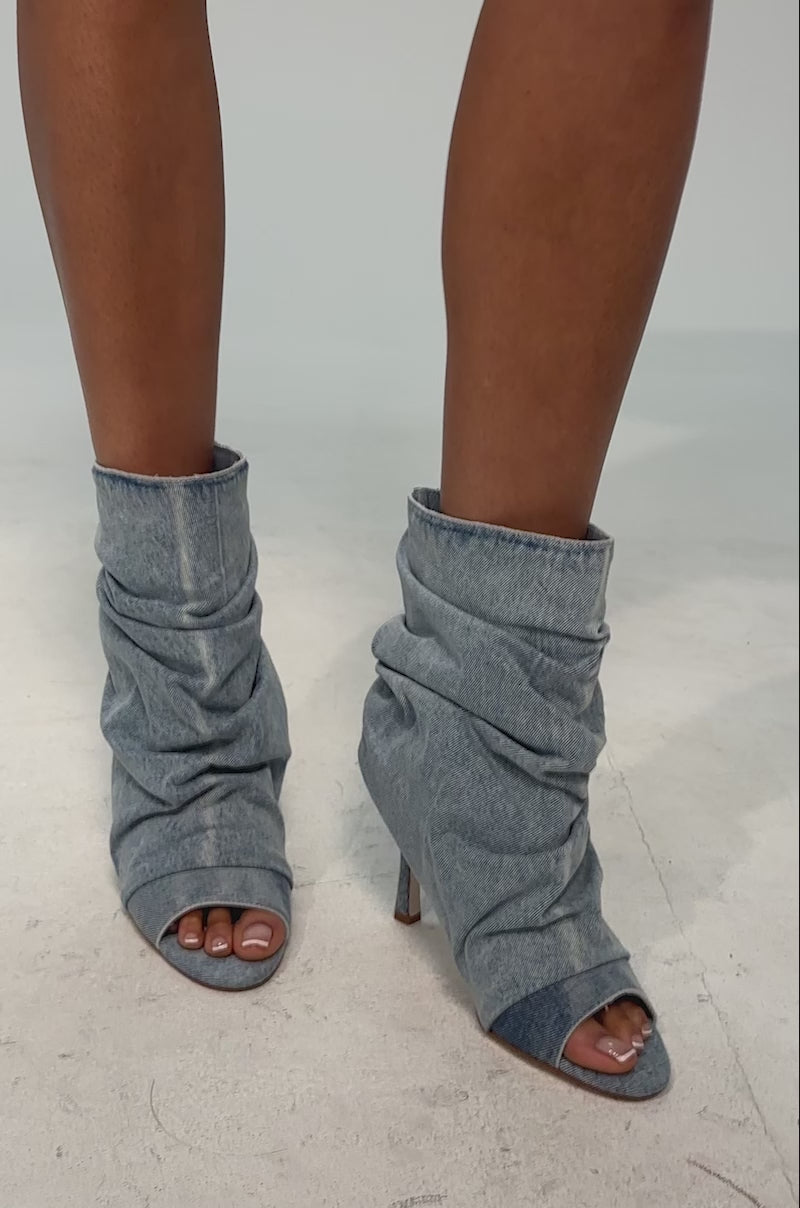 A video of a model wearing the AZALEA WANG APEX DENIM RUCHED STILETTO BOOTIE which features a light blue, slouchy denim design with ruched detailing, pointed toe, and stiletto heel.