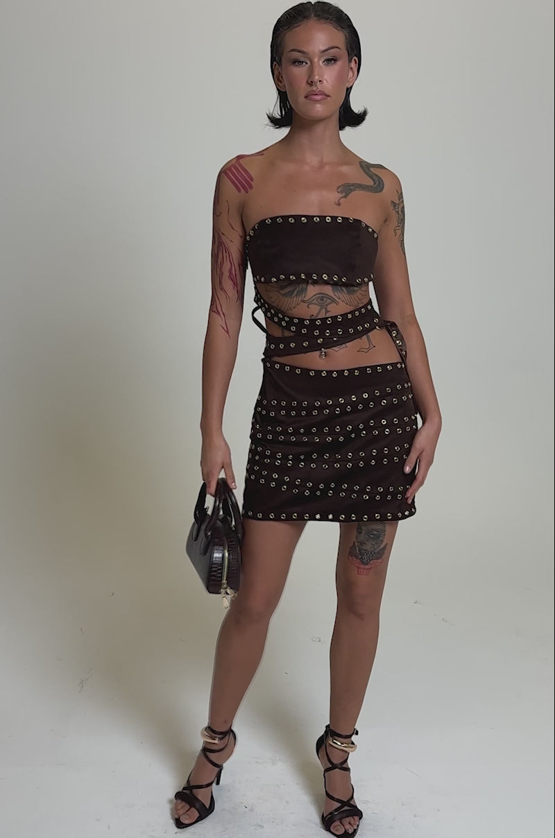 A video of a woman in the PARTY'S HERE SUEDE CUTOUT MINI DRESS, featuring brown suede with cutouts and gold grommet details. She pairs it with strappy brown heels, a dark handbag, visible tattoos, and wet-look hair.