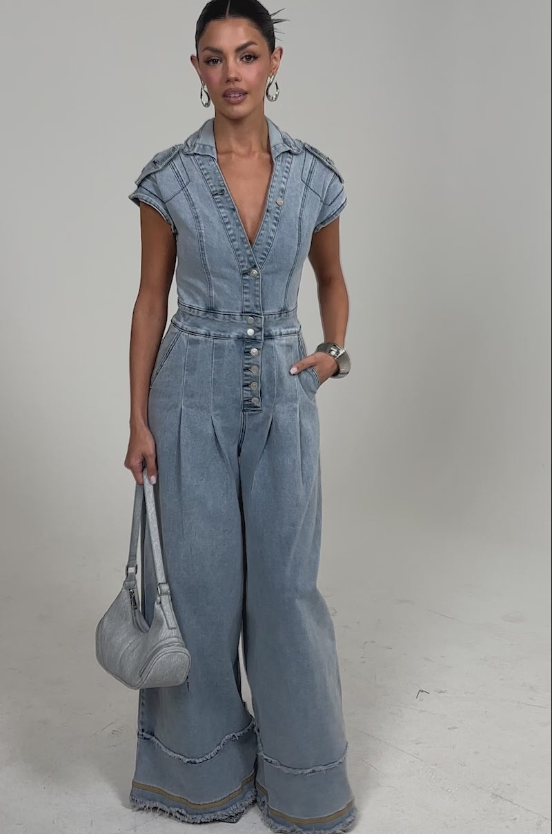 Video of woman modeling the AT LEAST I LOOK CUTE DENIM JUMPSUIT, featuring light-wash denim, short sleeves, deep V-neck, wide-leg pants, and silver buttons. She pairs it with a small gray purse, hoop earrings, and a bangle.