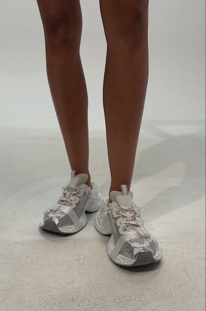 Video of woman modeling the AKIRA GOLD LOCKET WHITE SLIP ON SNEAKER featuring a white and gray design, open heel, mesh panels, chunky platform sole, and deconstructed details like loose fabric strips and exposed stitching.