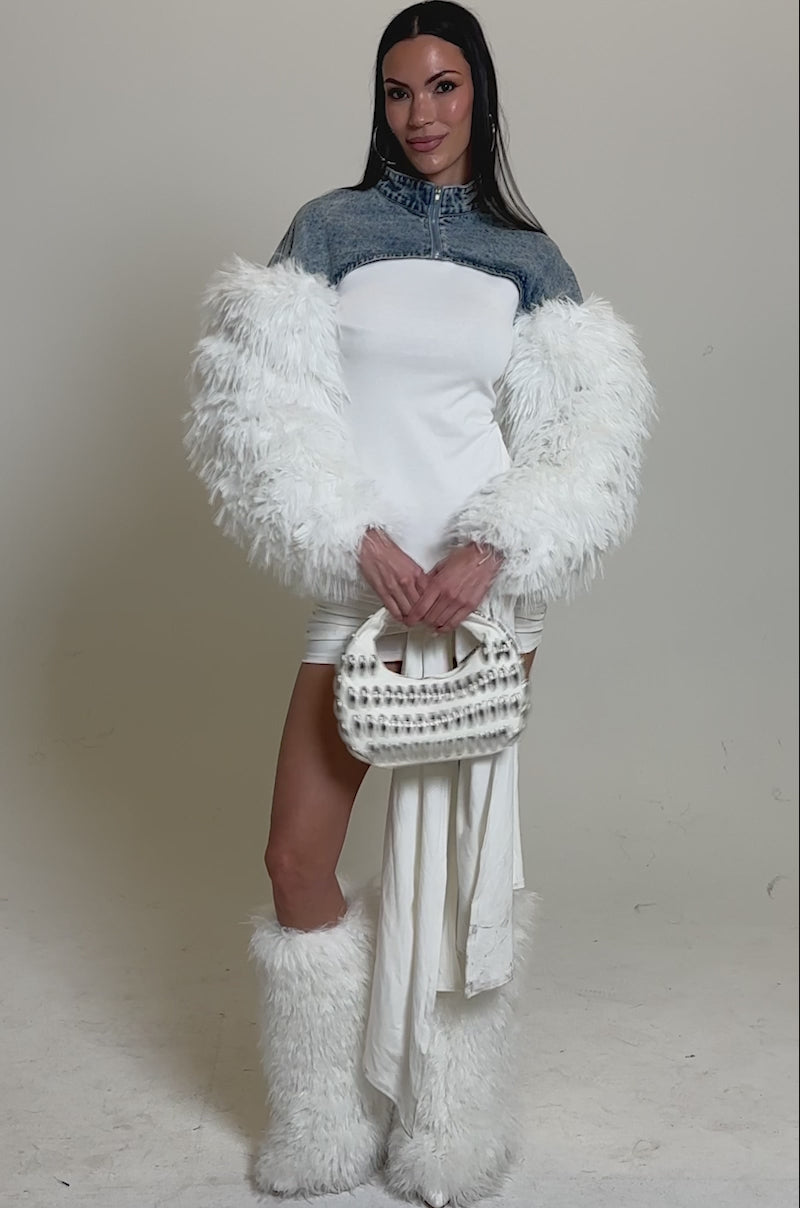 A video of a woman in a white dress with a ruched skirt, straight dark hair, and holds a studded handbag. She layers her look with the CELESTE FUR SLEEVE BOLERO and matching furry boots against a plain background.