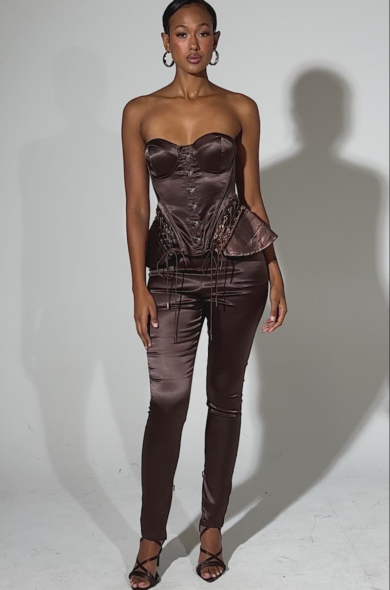 A video of a woman in the AINT MY PROBLEM CORSET, a shiny chocolate brown satin top with lace-up detailing, paired with matching fitted pants. She wears hoop earrings, strappy heels, and her hair is pulled back against a plain light background.