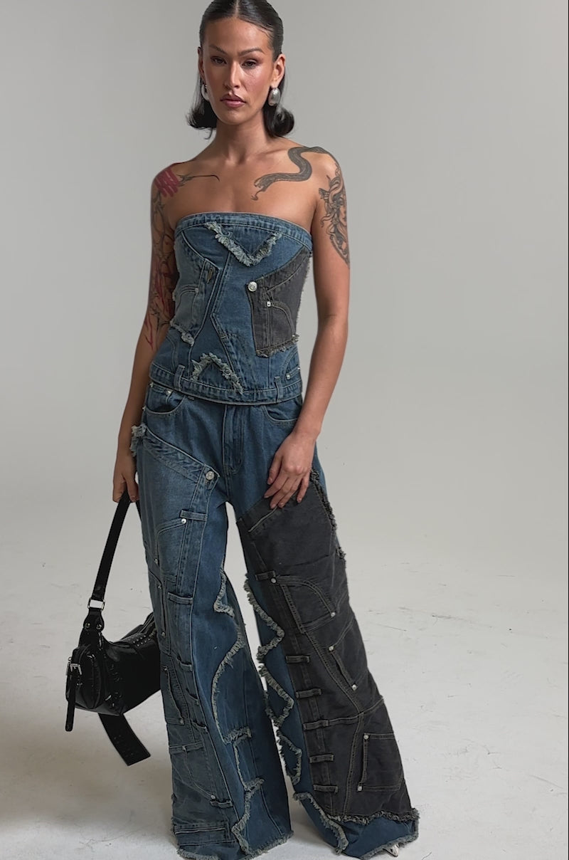 A video of a woman in the MIXED MATCH DENIM CORSET and wide-leg patchwork jeans, holding a black handbag against a plain backdrop.
