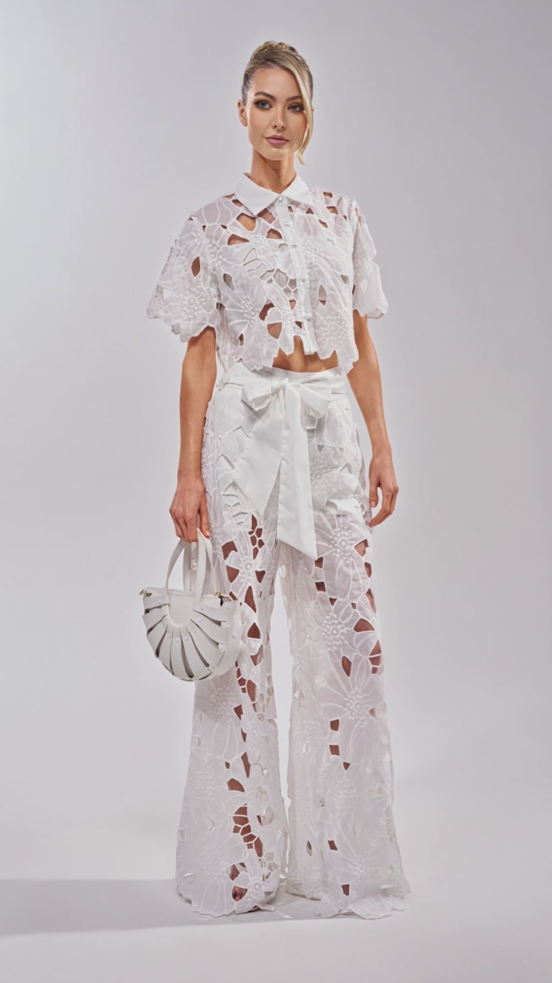 Video of woman modeling the DOLLY FLORAL PALAZZO PANT IN WHITE, which features a high waist and large floral cut-outs, paired with an oversized front bow and a small white handbag. 