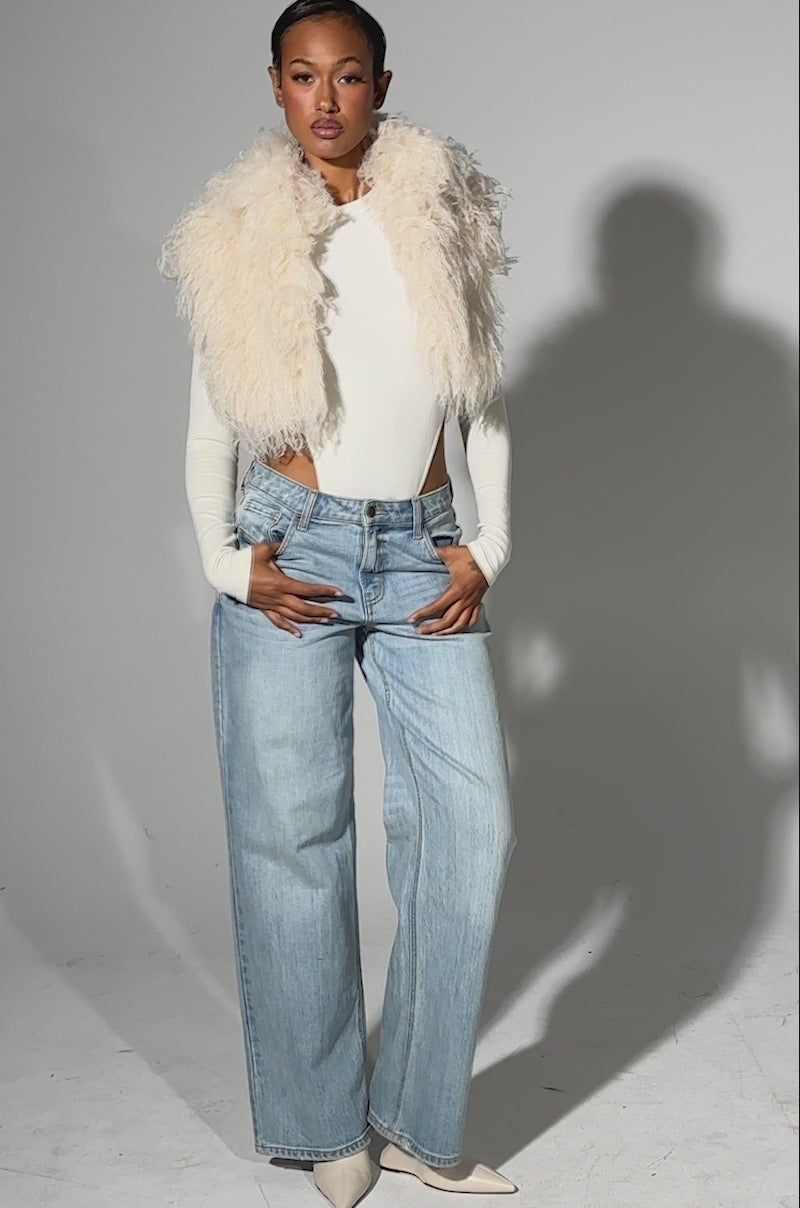 A video of a person standing against a plain background wearing the COCO CROPPED FUR VEST IN BEIGE over a fitted white top, paired with light blue high-waisted wide-leg jeans and pointed white shoes.