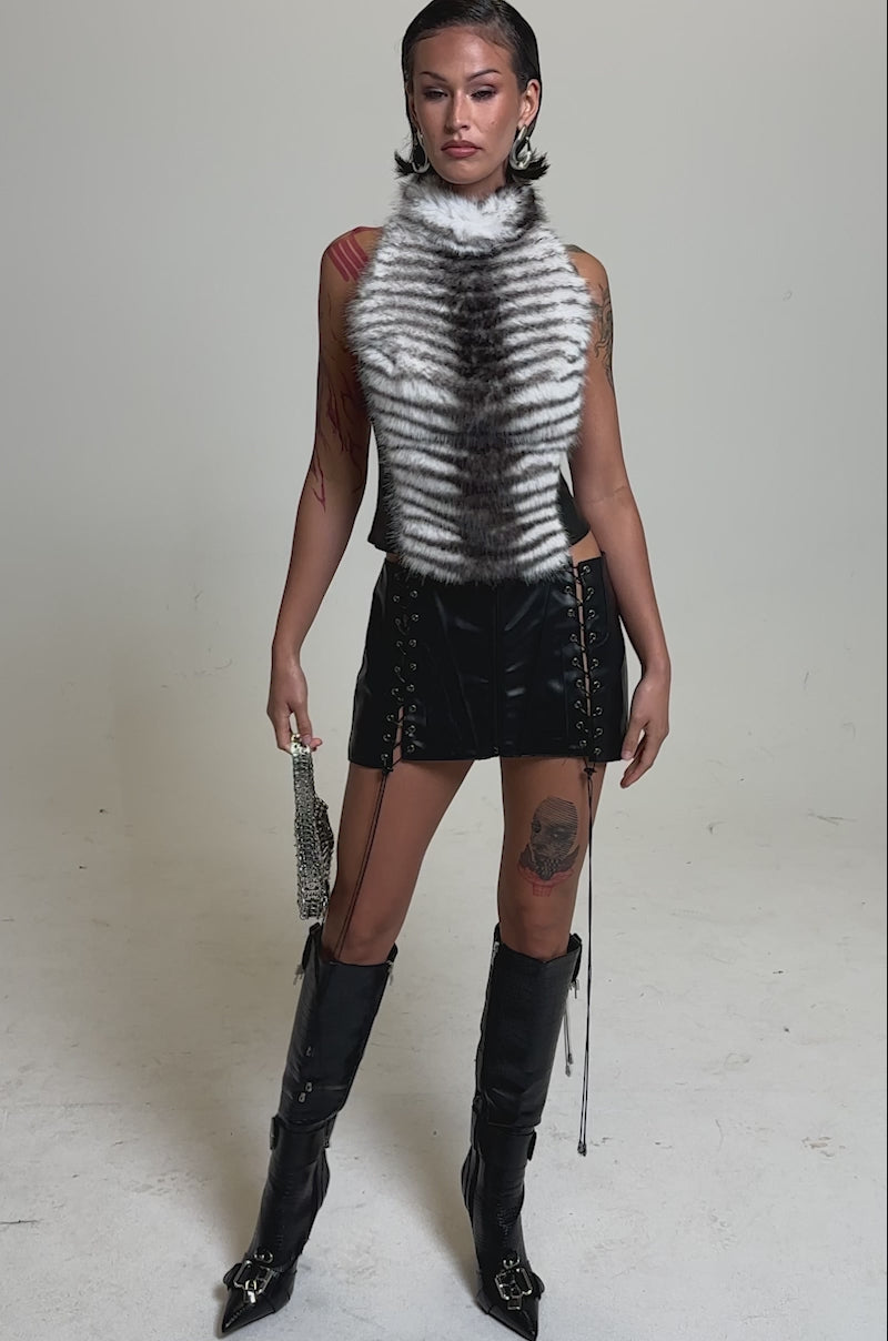 A video of a model in the I'M MY OWN SPIRIT ANIMAL MOCK NECK FUR TOP, paired with a black faux leather mini skirt with lace-up sides, tall black boots, and a silver handbag. 