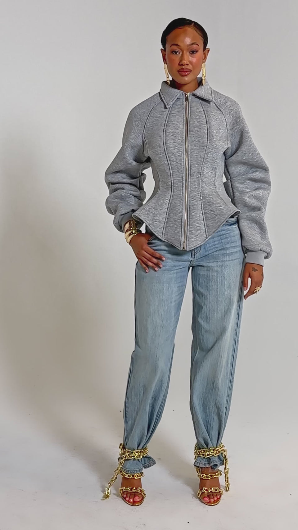 A video of a woman wearing the MONEY TALKS NEOPRENE JACKET with puffed sleeves, light blue flared jeans, gold heels, and jewelry against a plain white background.