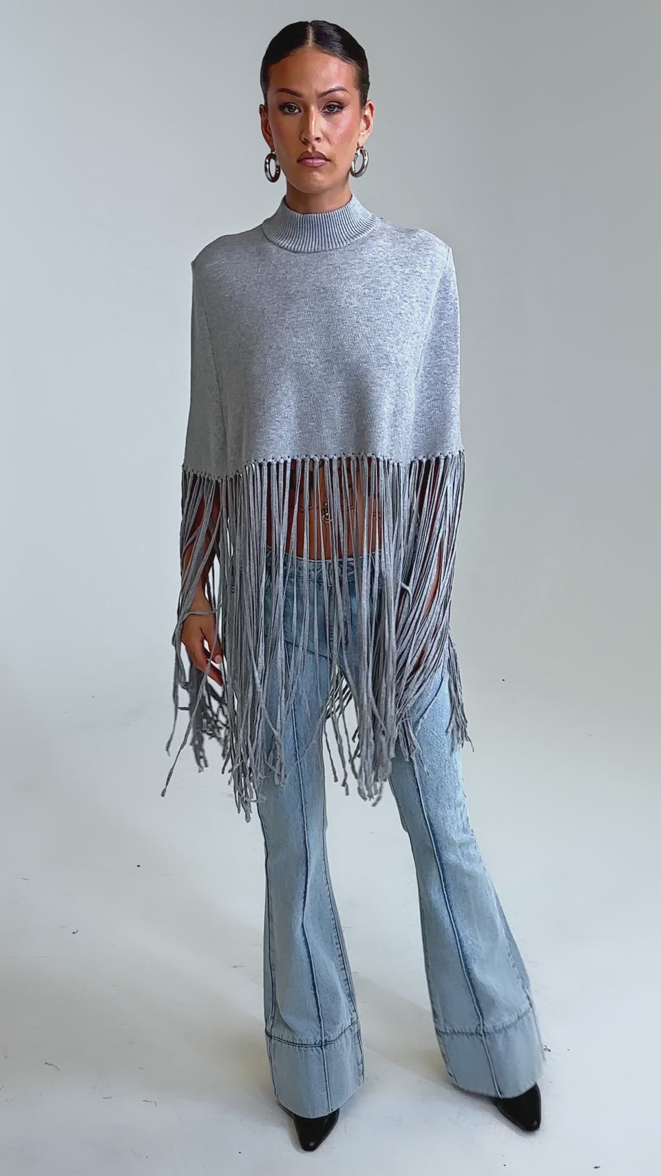 Video of woman modeling the FRINGE FOR DAYS SWEATER, a gray high-neck poncho with long, thick fringe, styled over blue jeans. She stands confidently against a plain background, highlighting this chic knit layering piece.