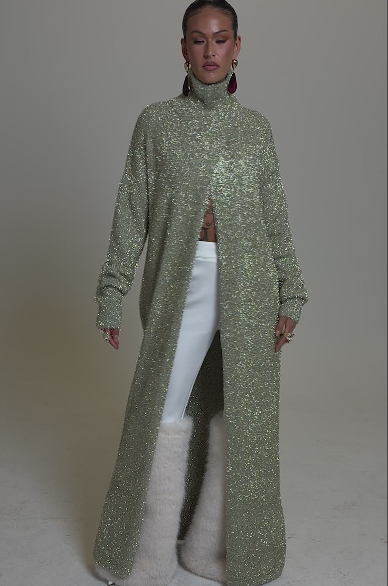 A video of a model wearing the WORTHY OF LOVE LONG SLEEVE METALLIC SWEATER IN GOLD under a sparkly open-front coat and white high-waisted pants with pale fur at the lower legs, accessorized with bold earrings and showcasing visible torso tattoos.
