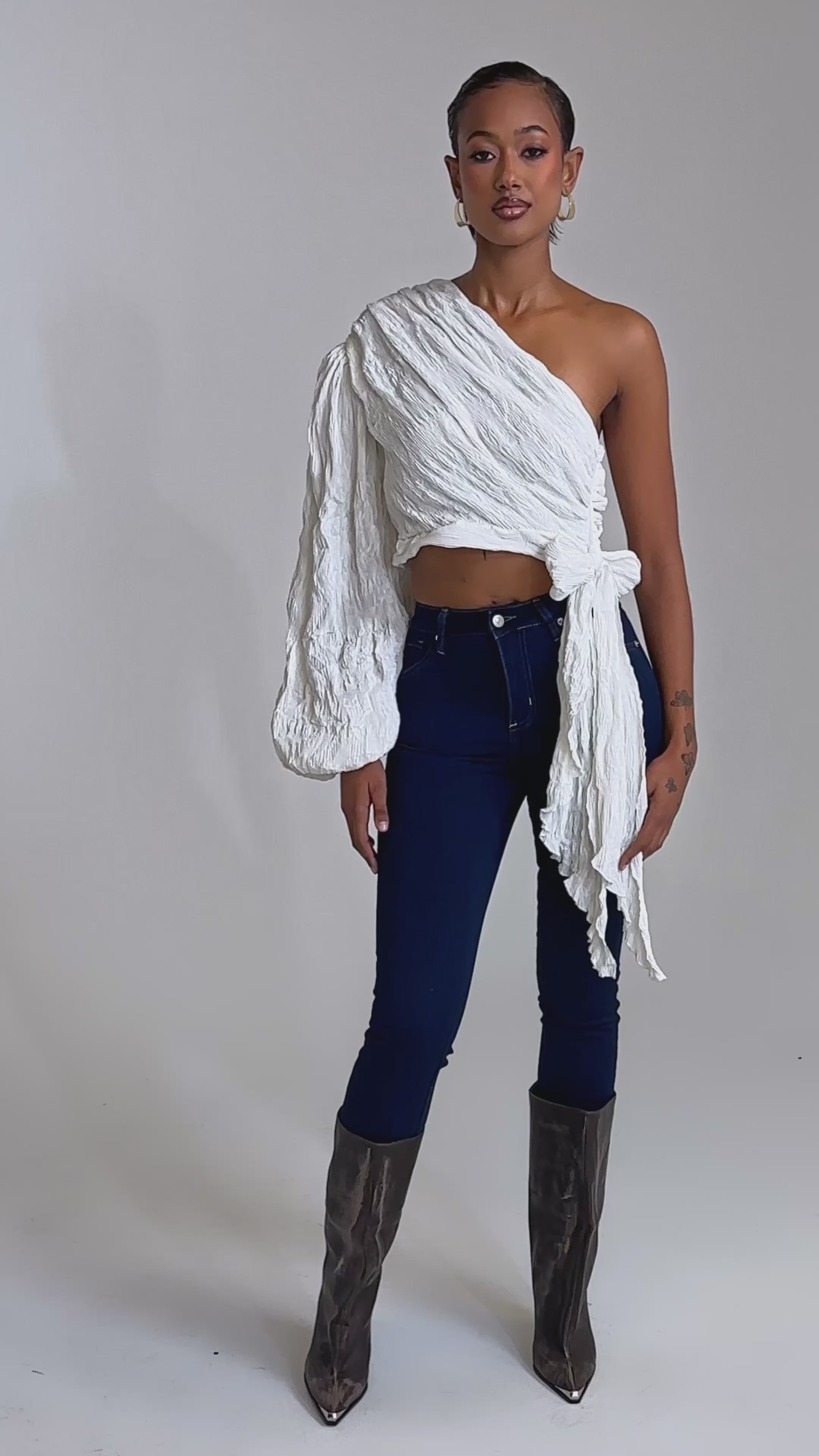 A video of a woman wearing the GROOVY BABY SIDE TIE BLOUSE—a white, one-shoulder cropped top—paired with dark blue skinny jeans and knee-high gray boots.