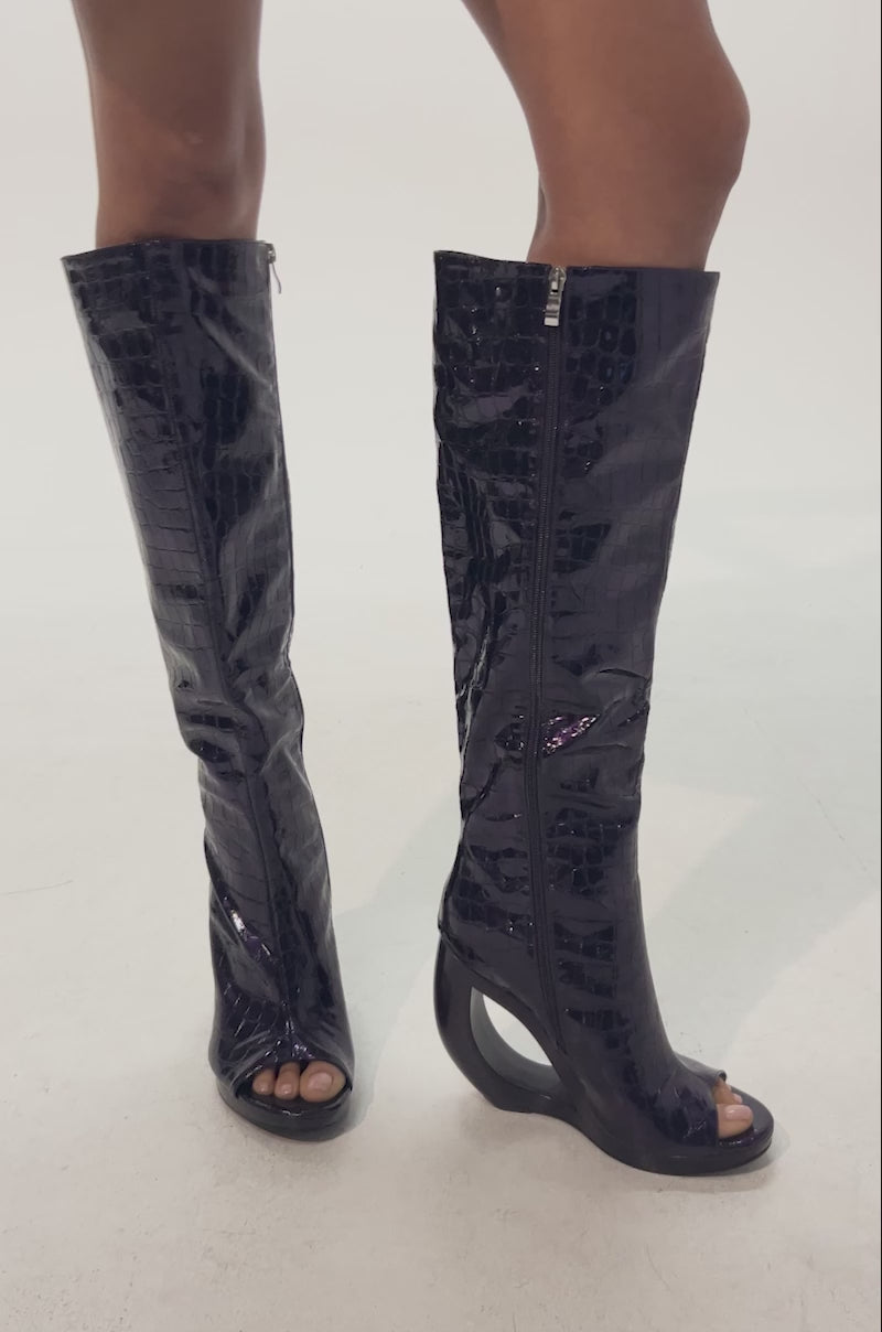 Video of woman modeling the AZALEA WANG CAMILLA PURPLE NOIR CROC OPEN TOE WEDGE BOOTS—knee-high, shiny purple boots featuring croc-embossed texture, a unique open sculptural wedge heel, and a peep-toe design.