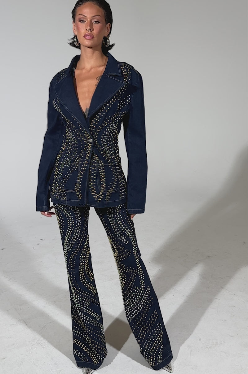 A video of a person wearing CHEYENNE STUDDED FLARE DENIM featuring gold studs and swirling silver and gold accents, paired with a dark blue fitted blazer.