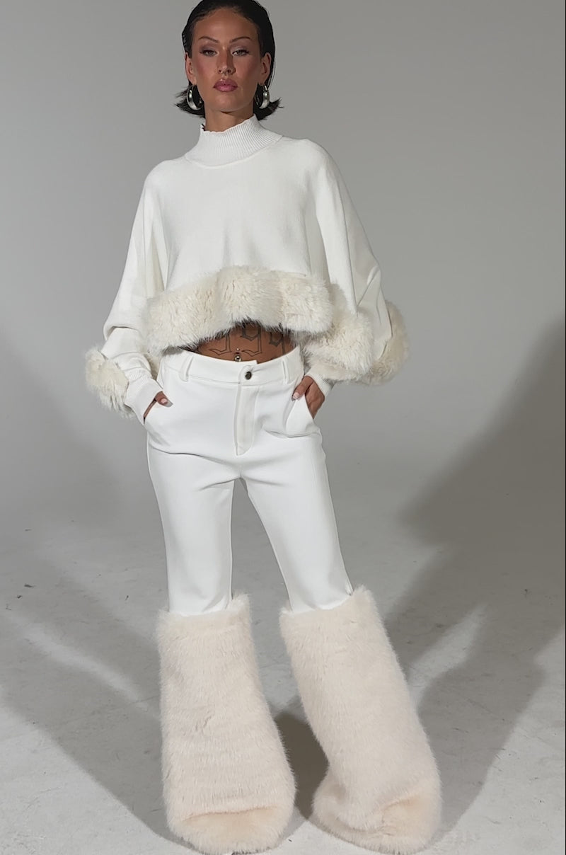 A video of a person modeling the QUIET LUX FUR TRIM SWEATER IN WHITE, a cropped poncho-style sweater with fluffy fur trim, paired with high-waisted white pants and oversized furry leg warmers against a light background.