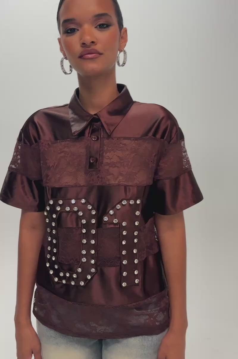 Video of a woman modeling the NEW AUTHORITY LACE SHORT SLEEVE POLO—a lustrous burgundy shirt featuring sheer floral lace accents and a “01” motif outlined in metal studs.